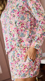 A woman wearing a matching two-piece pajama set with a pink, blue, and green floral pattern, standing in a bedroom next to a window and vanity.