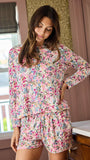 A woman wearing a matching long-sleeve top and shorts pajama set with a colorful floral print, standing in a bedroom setting.