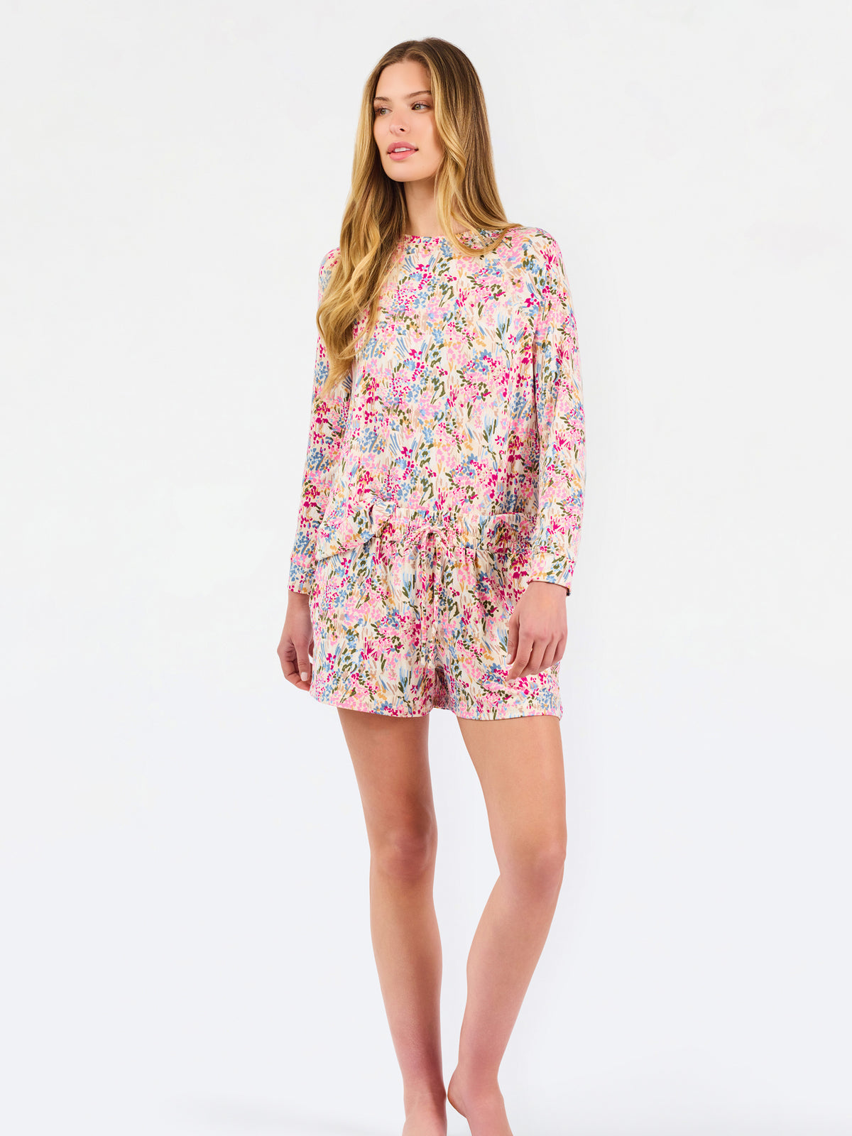 Blonde long haired woman wearing a multi colored floral long sleeve and shorts pajama set on a white background