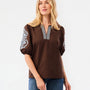 A woman modeling a brown short-sleeve top with white embroidered puff sleeves and a silver and white embroidered V-neckline, paired with blue jeans against a white studio background.