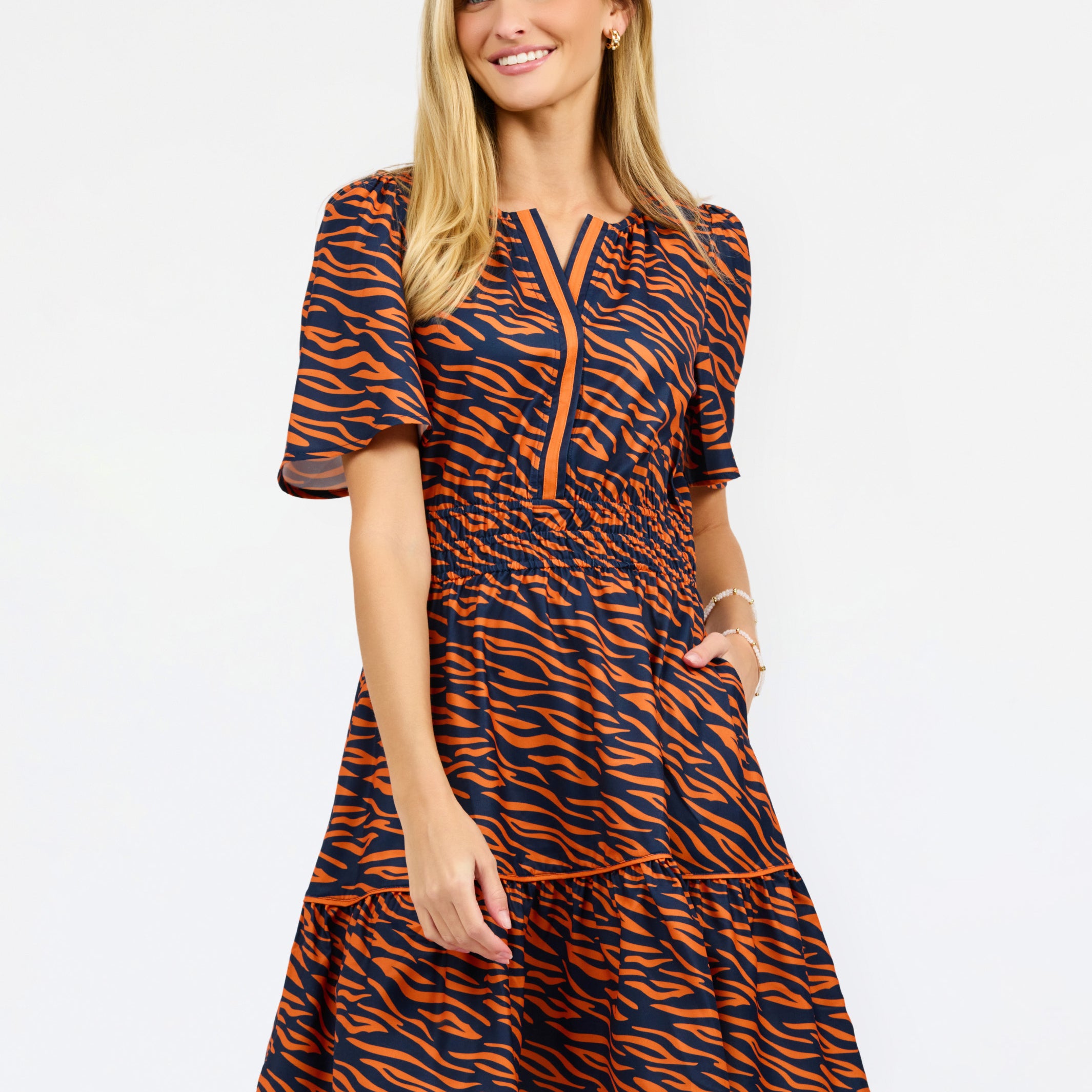 A woman modeling a short-sleeve tiered dress with an orange and navy tiger stripe pattern, featuring a split neckline with orange piping details, set against a plain white studio background.