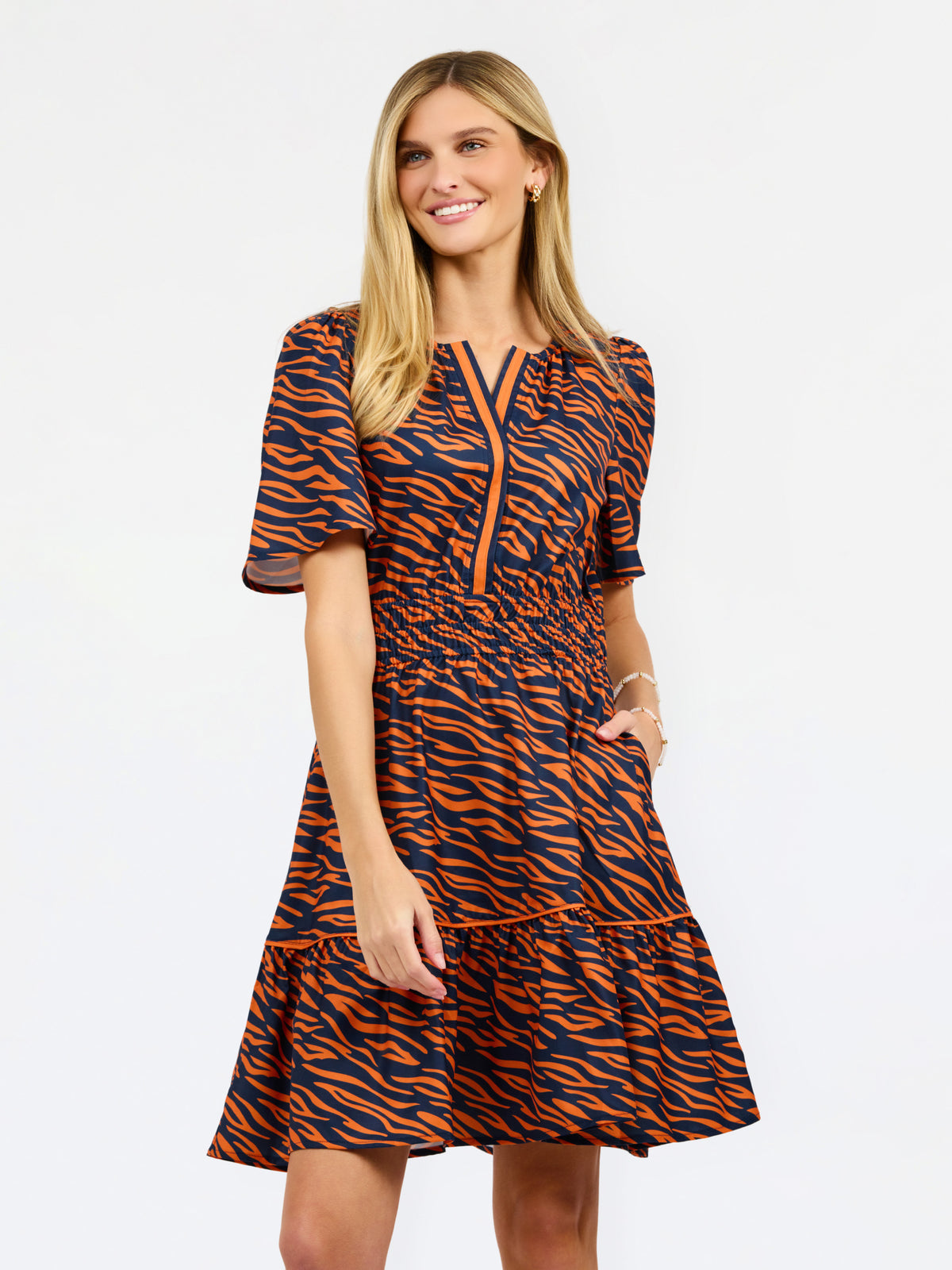 A woman modeling a short-sleeve tiered dress with an orange and navy tiger stripe pattern, featuring a split neckline with orange piping details, set against a plain white studio background.