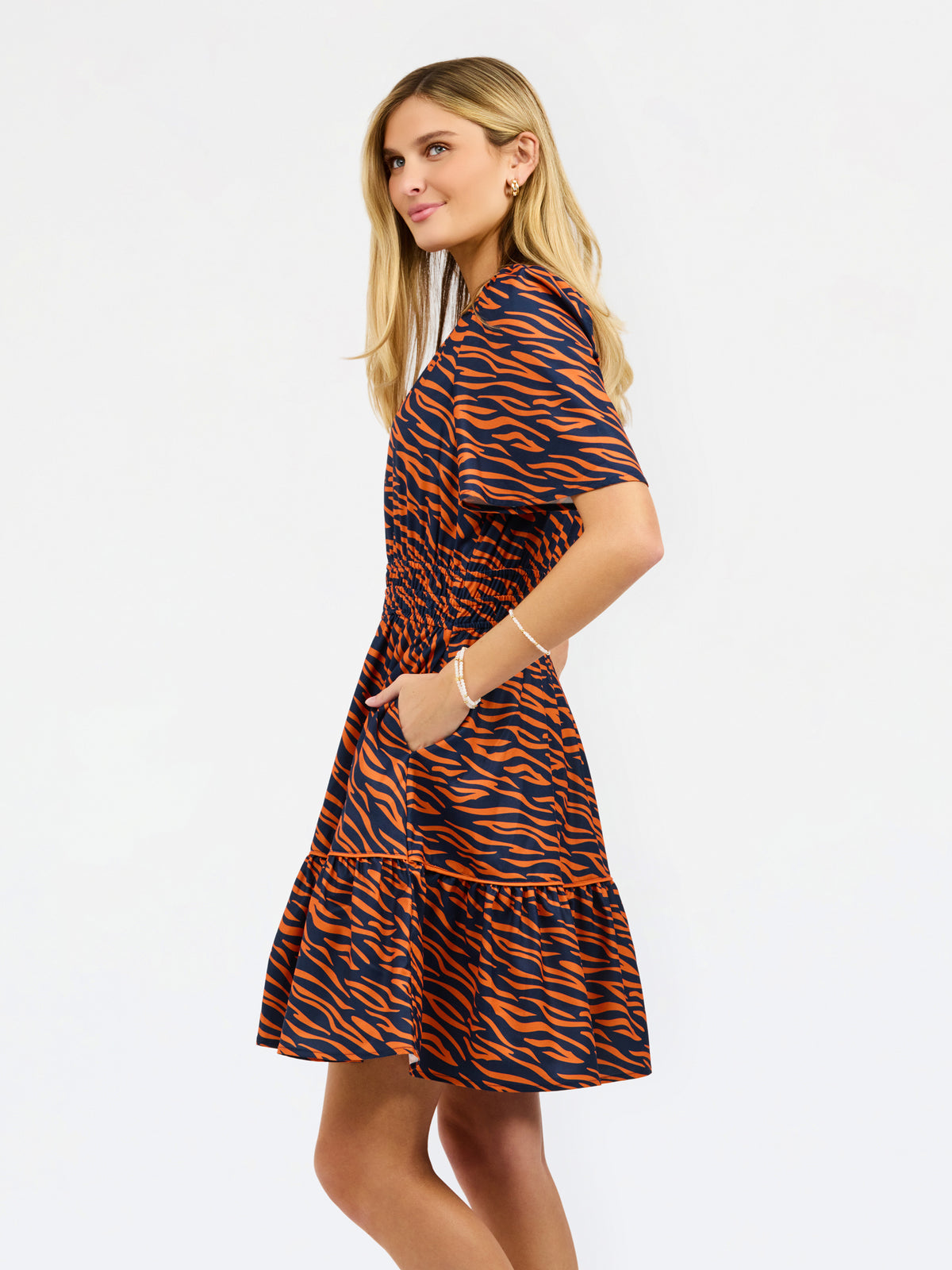A woman modeling a short-sleeve mini dress with a bold orange and navy tiger stripe pattern, shown from a side-back angle against a plain white studio background.