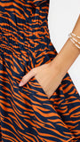 Close-up of a sleeveless dress with a bold orange and navy blue tiger stripe pattern, featuring a smocked elastic waist and a side pocket with a hand inserted. The model's arm is adorned with double-strand gold beaded bracelets against a clean white background.