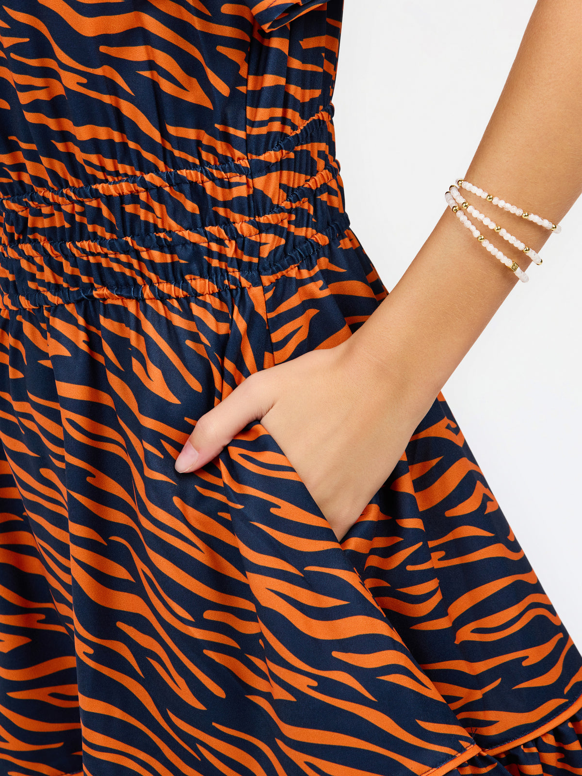 Close-up of a sleeveless dress with a bold orange and navy blue tiger stripe pattern, featuring a smocked elastic waist and a side pocket with a hand inserted. The model's arm is adorned with double-strand gold beaded bracelets against a clean white background.