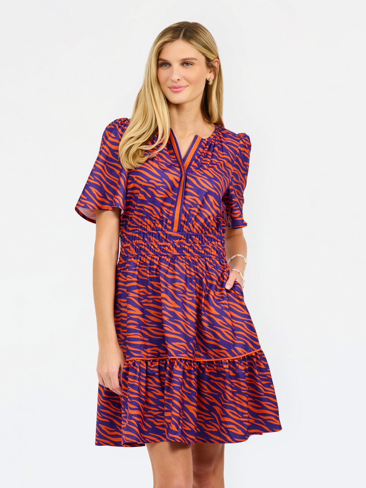 A woman modeling a short-sleeve tiered dress with a purple and orange tiger-like pattern, featuring a split neckline with orange trim and side pockets, set against a plain white background.