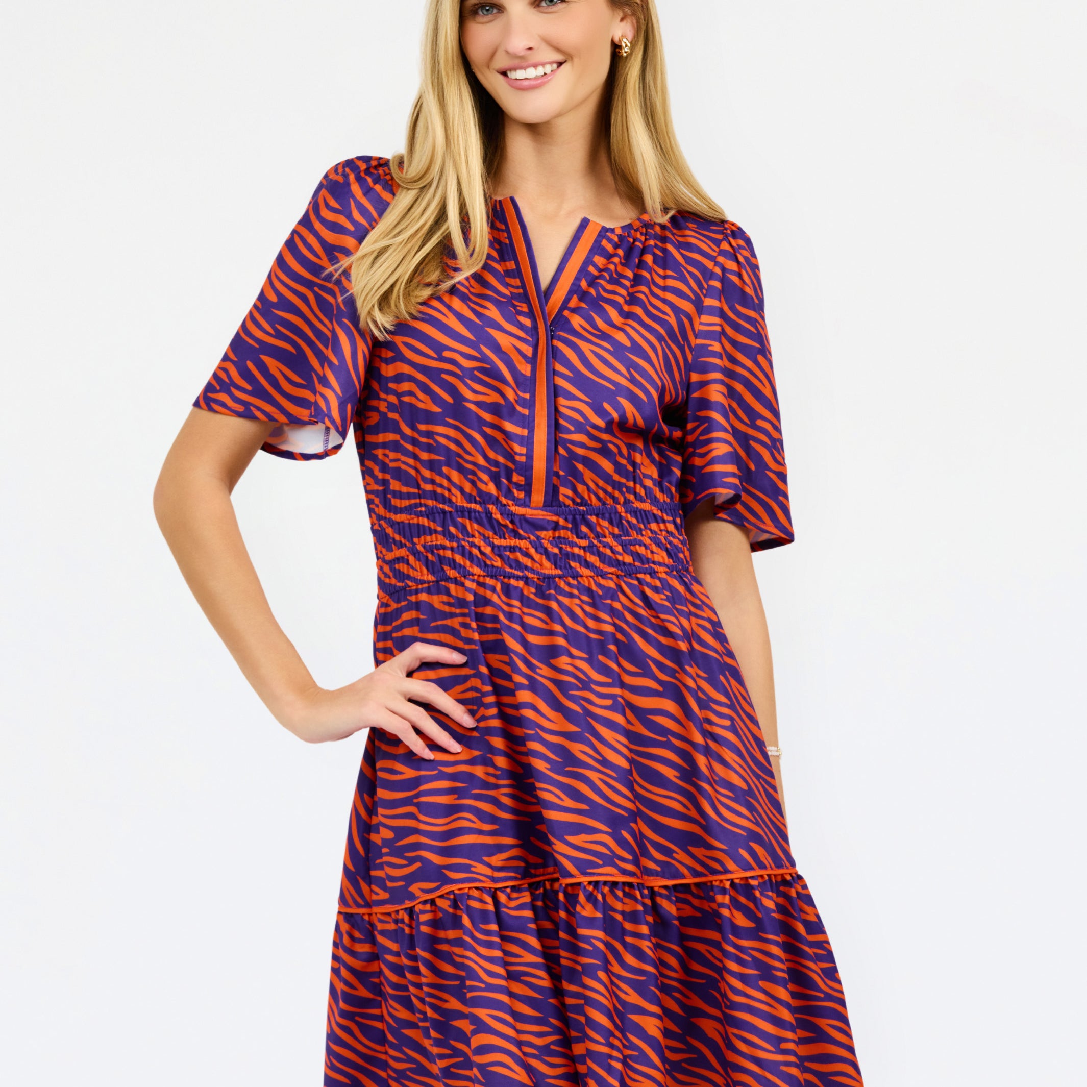 A woman modeling a short-sleeve tiered dress with a purple and orange tiger-like pattern, featuring a split neckline with orange trim and side pockets, set against a plain white background.
