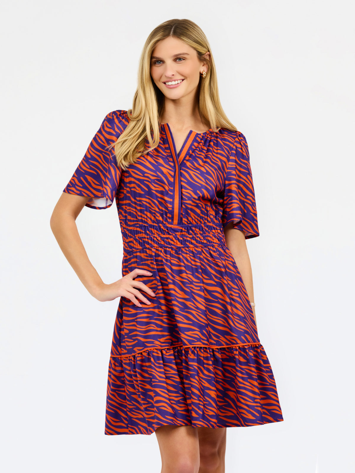 A woman modeling a short-sleeve tiered dress with a purple and orange tiger-like pattern, featuring a split neckline with orange trim and side pockets, set against a plain white background.