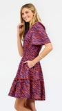 A woman modeling a short-sleeve tiered dress with a purple and orange tiger-like pattern, featuring a split neckline with orange trim and side pockets, set against a plain white background.