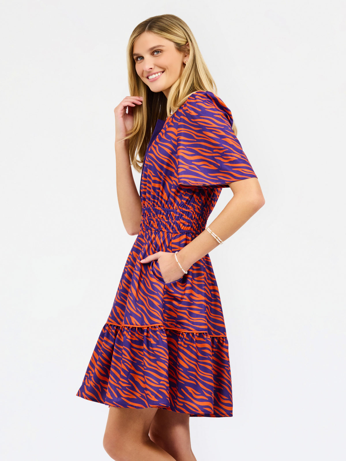 A woman modeling a short-sleeve tiered dress with a purple and orange tiger-like pattern, featuring a split neckline with orange trim and side pockets, set against a plain white background.