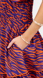 Close-up of a person wearing a tiered dress with an orange and purple wavy animal-style print, shown from the chest down with one hand in a side pocket. The person is also wearing two beaded bracelets.