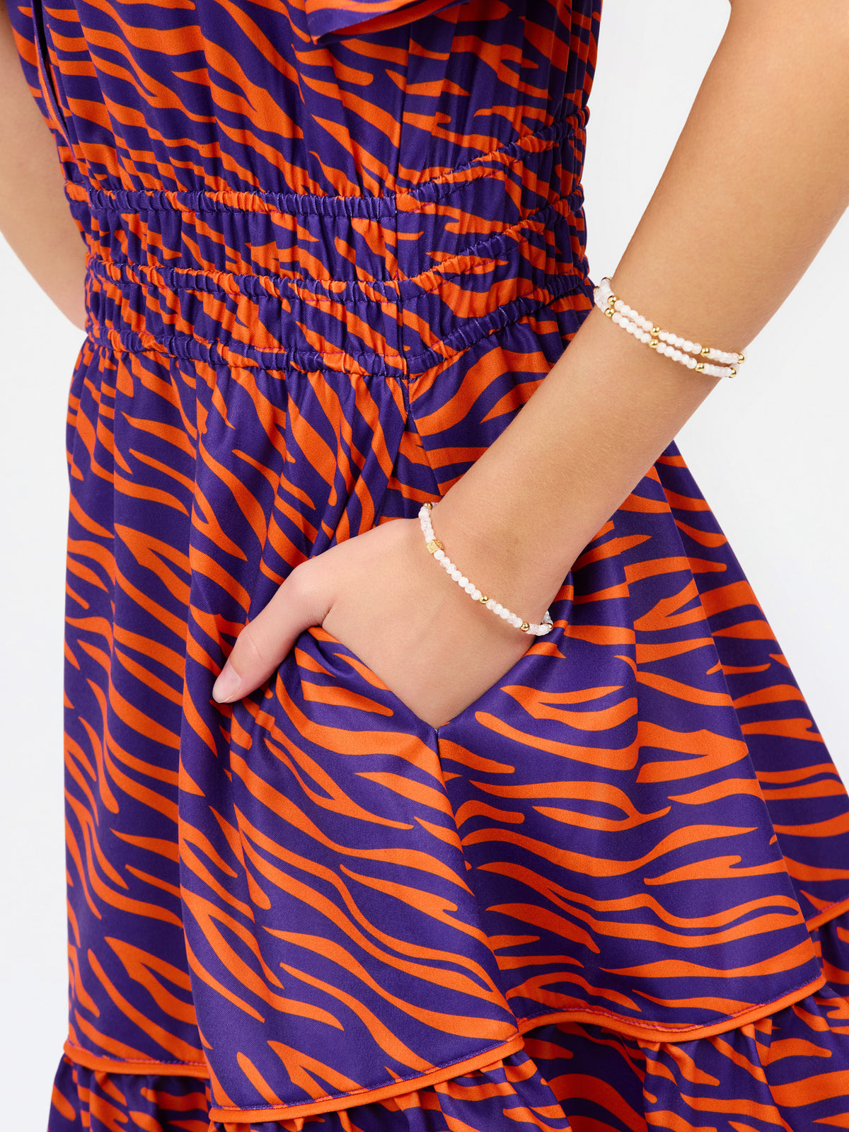Close-up of a person wearing a tiered dress with an orange and purple wavy animal-style print, shown from the chest down with one hand in a side pocket. The person is also wearing two beaded bracelets.