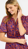 A woman modeling a short-sleeve tiered dress with a purple and orange tiger-like pattern, featuring a split neckline with orange trim and side pockets, set against a plain white background.