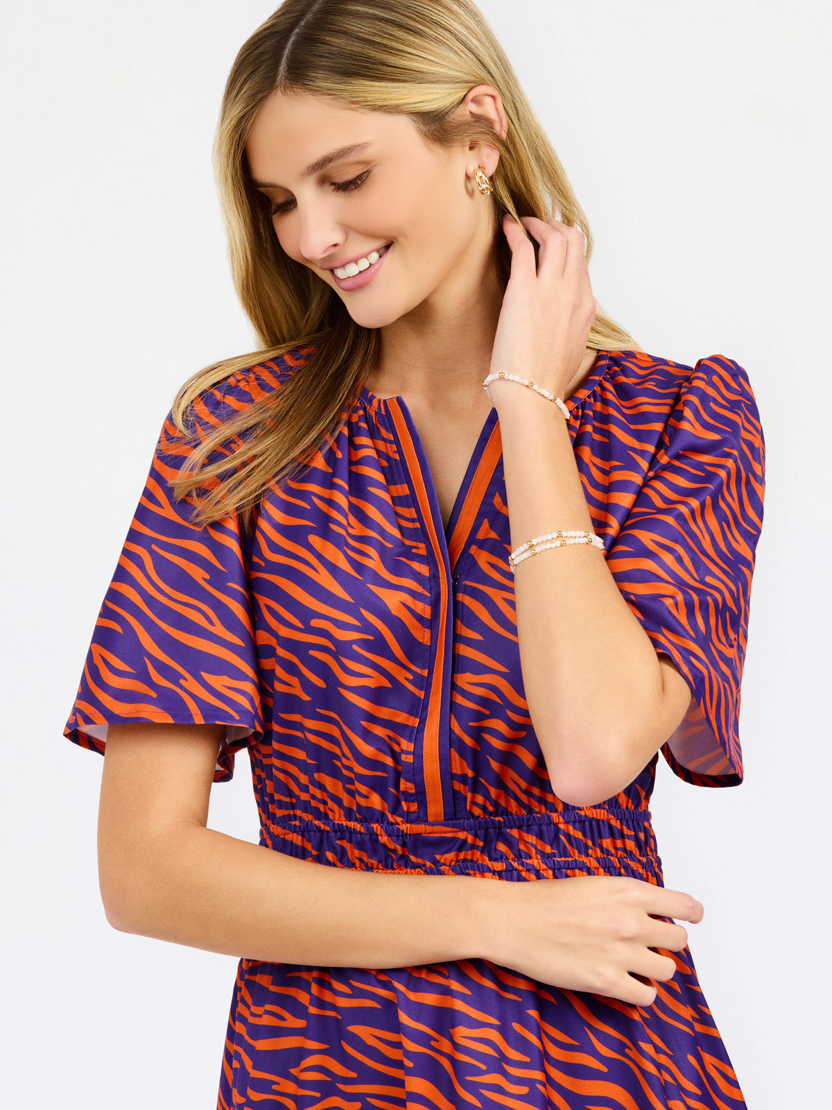 A woman modeling a short-sleeve tiered dress with a purple and orange tiger-like pattern, featuring a split neckline with orange trim and side pockets, set against a plain white background.