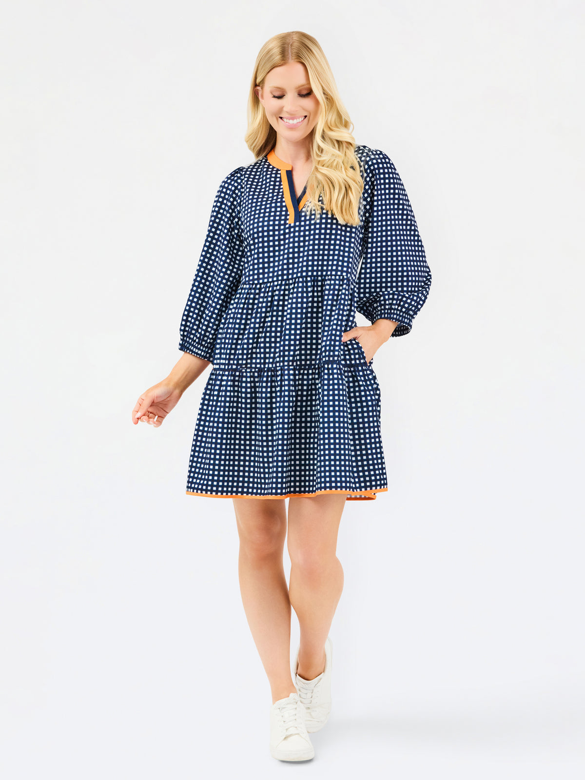 A woman modeling a navy blue and white gingham tiered dress with orange trim details on the neckline and hem, set against a plain light gray background.