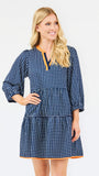 A woman modeling a navy blue and white gingham tiered dress with orange trim details on the neckline and hem, set against a plain light gray background.