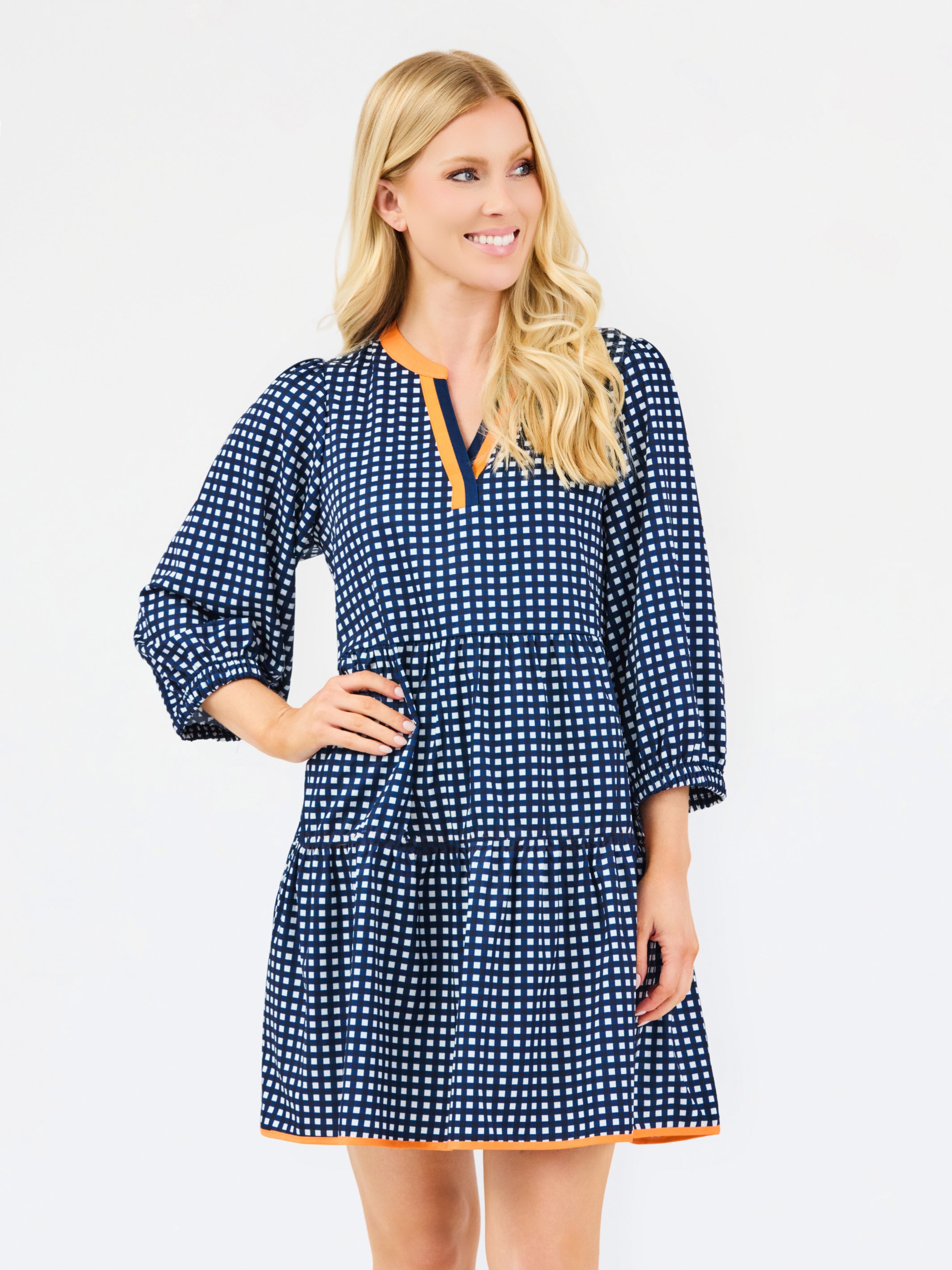 A woman modeling a navy blue and white gingham tiered dress with orange trim details on the neckline and hem, set against a plain light gray background.