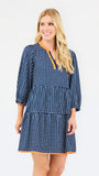 A woman modeling a navy blue and white gingham tiered dress with orange trim details on the neckline and hem, set against a plain light gray background.