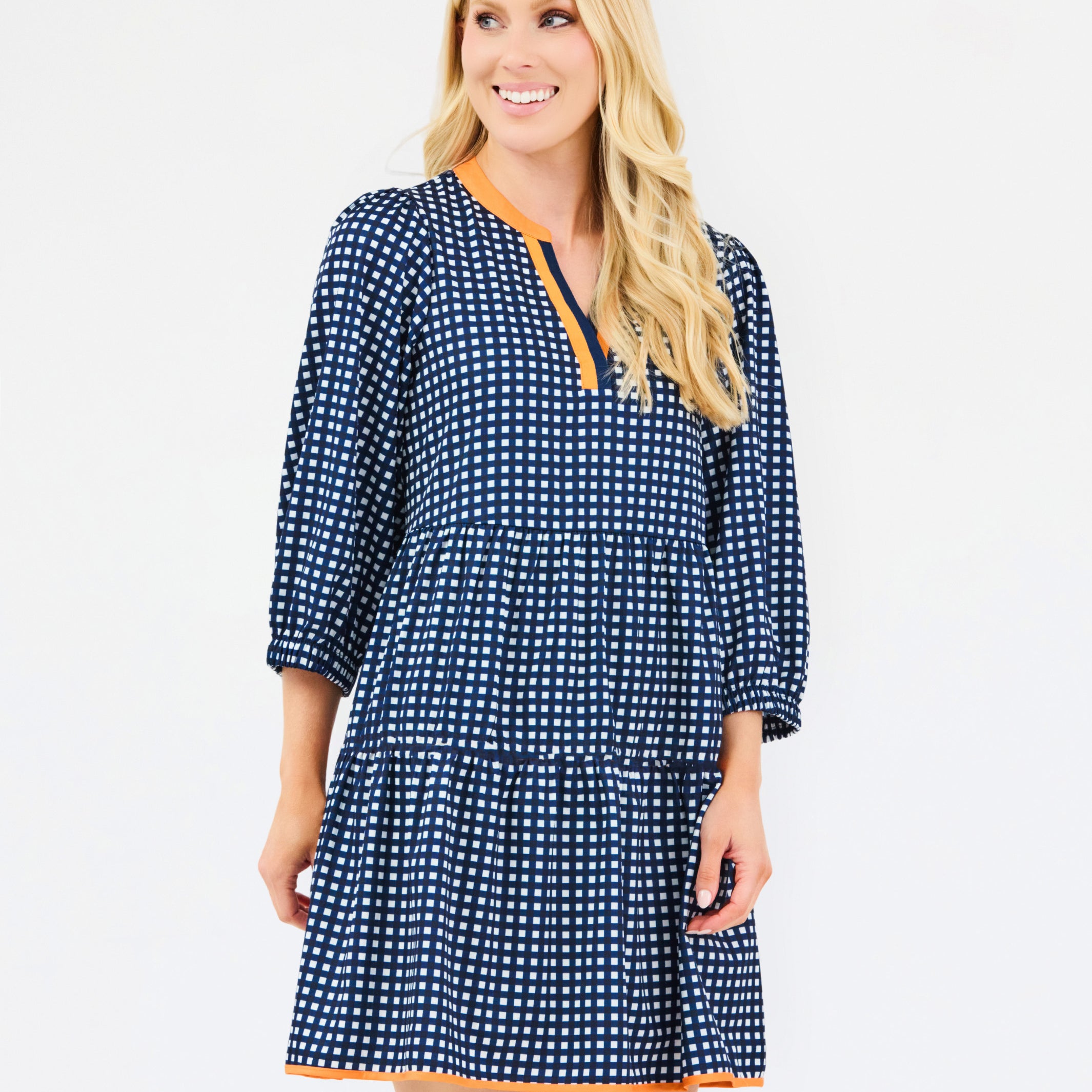 A woman modeling a navy blue and white gingham tiered dress with orange trim details on the neckline and hem, set against a plain light gray background.