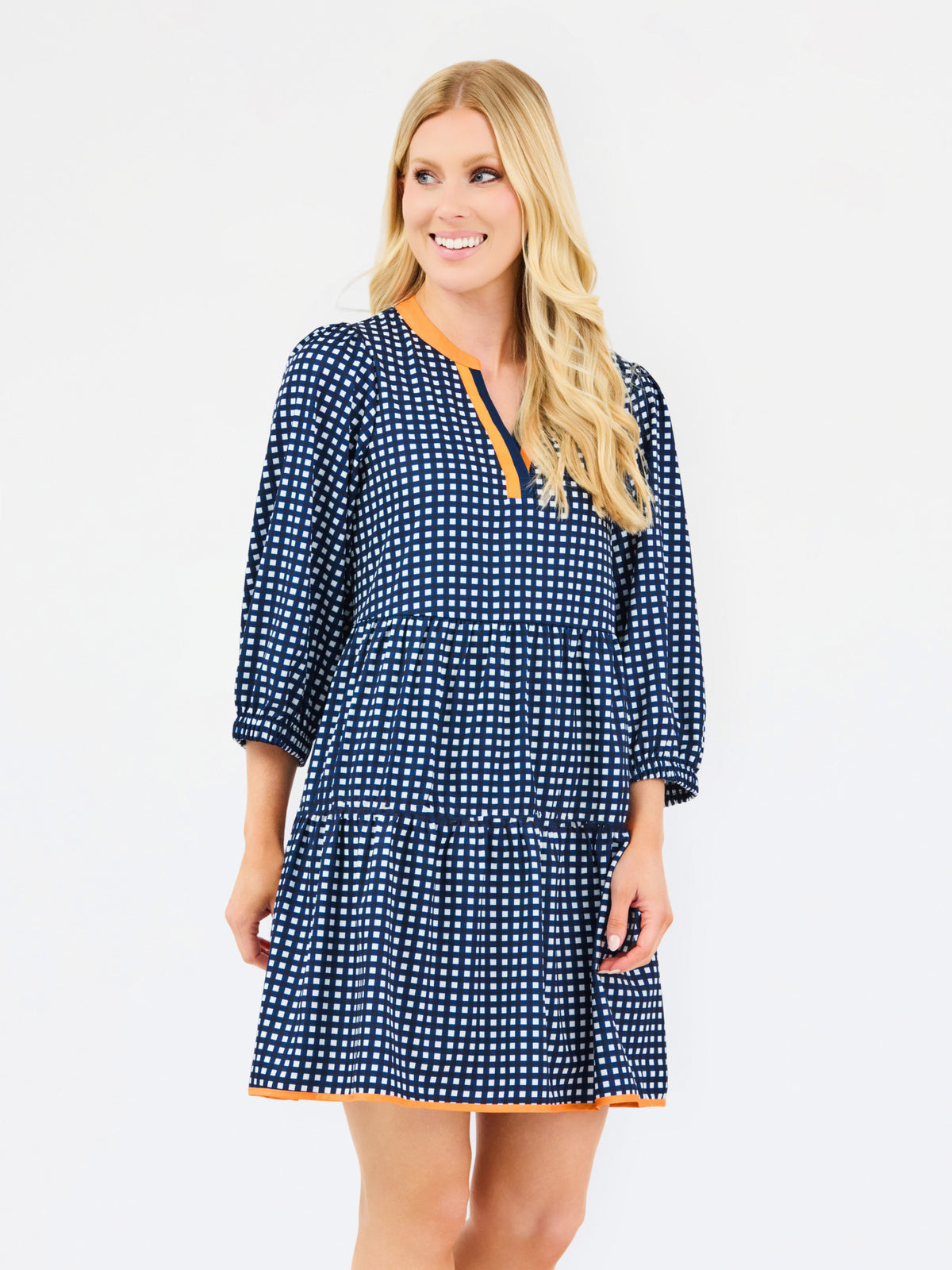 A woman modeling a navy blue and white gingham tiered dress with orange trim details on the neckline and hem, set against a plain light gray background.