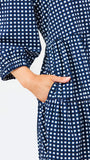 A woman modeling a navy blue and white gingham tiered dress with orange trim details on the neckline and hem, set against a plain light gray background.