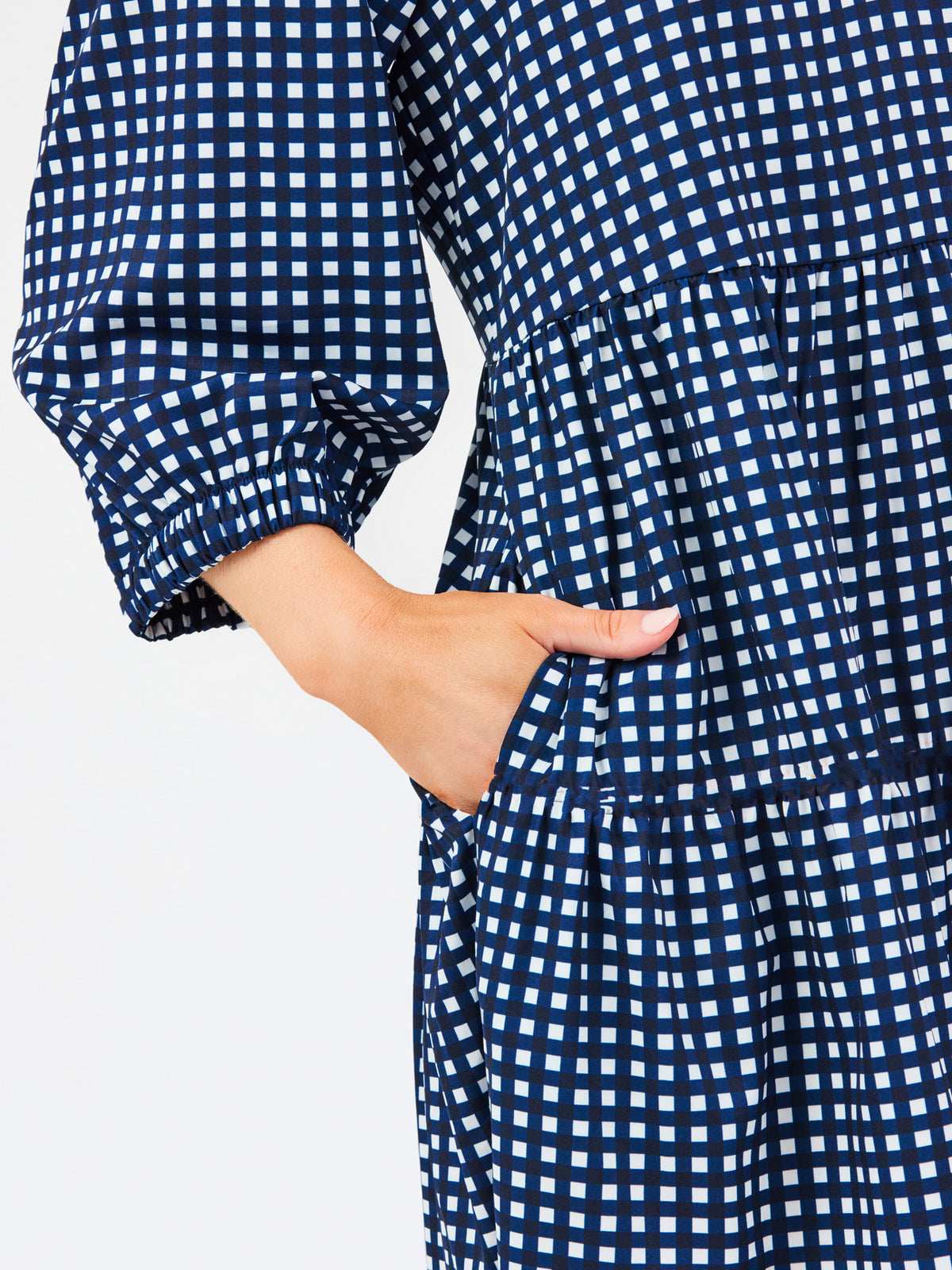 A woman modeling a navy blue and white gingham tiered dress with orange trim details on the neckline and hem, set against a plain light gray background.