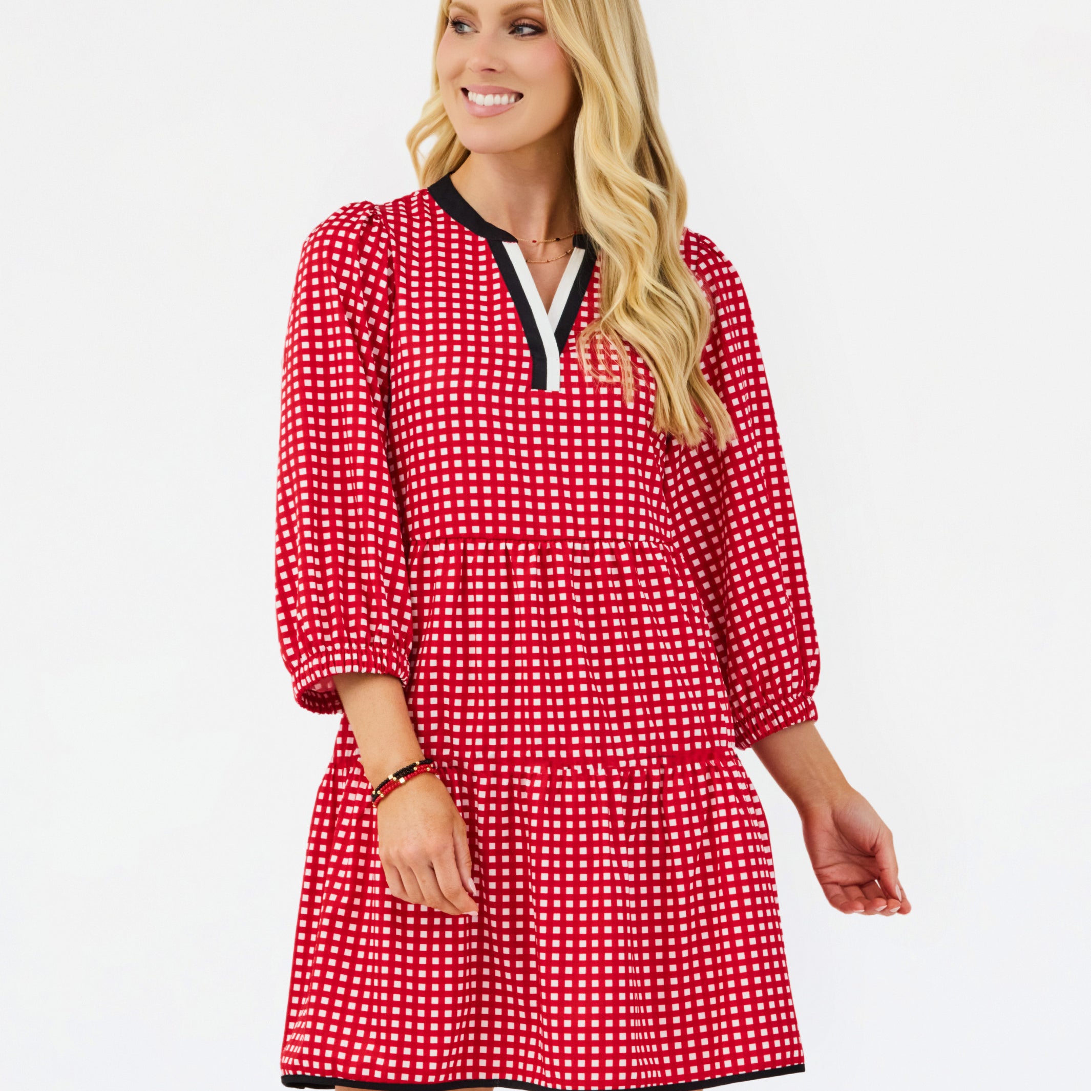 A woman modeling a red and white gingham tiered dress with black trim on the collar and hem, set against a plain white studio background.