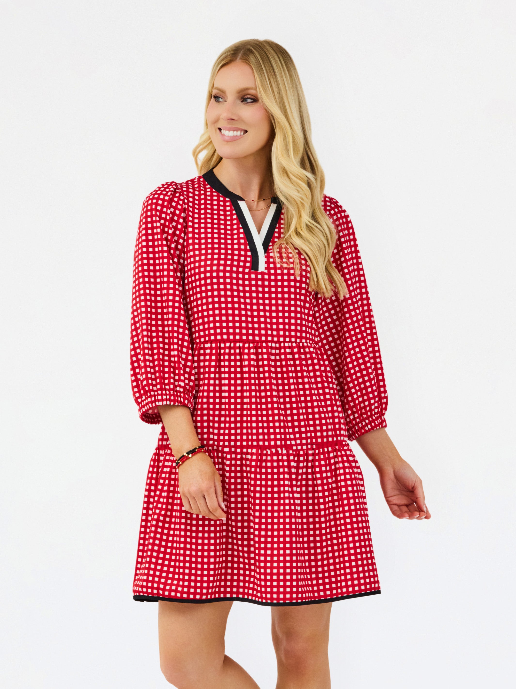 A woman modeling a red and white gingham tiered dress with black trim on the collar and hem, set against a plain white studio background.