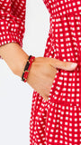 A close-up of a person wearing a red and white gingham dress, with one hand tucked into a pocket and the other showing a beaded bracelet.