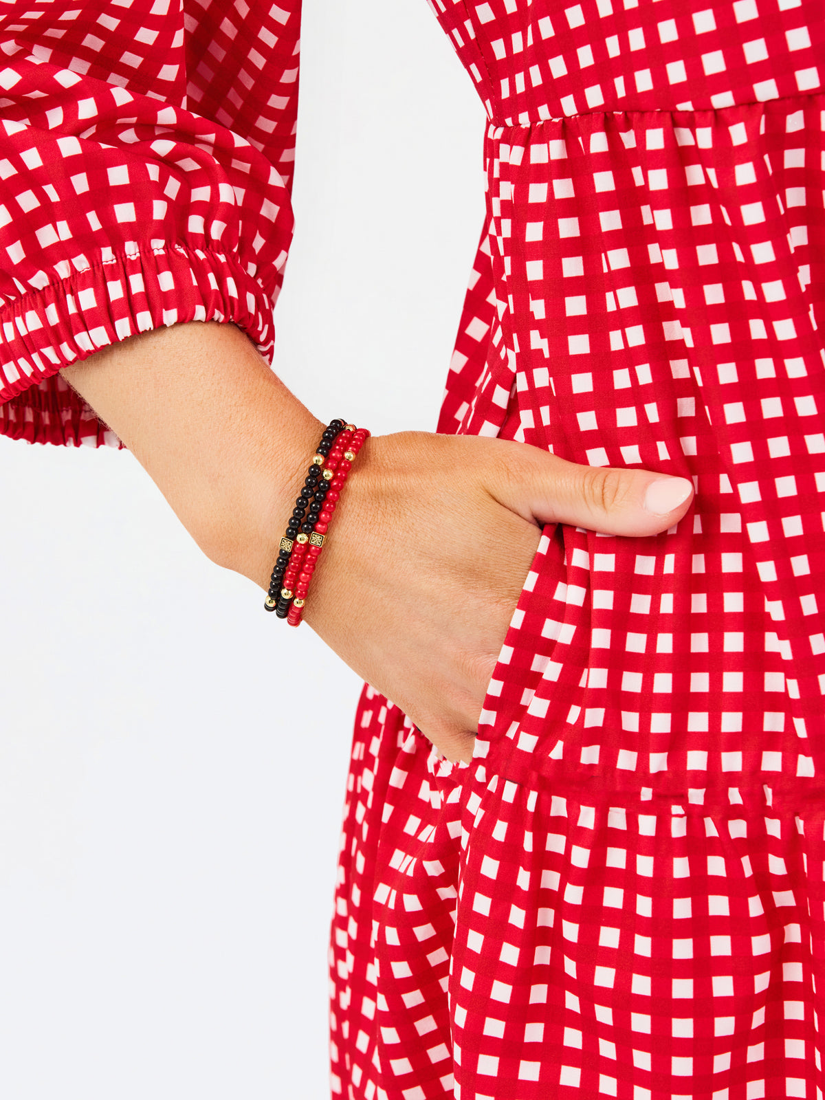 A close-up of a person wearing a red and white gingham dress, with one hand tucked into a pocket and the other showing a beaded bracelet.