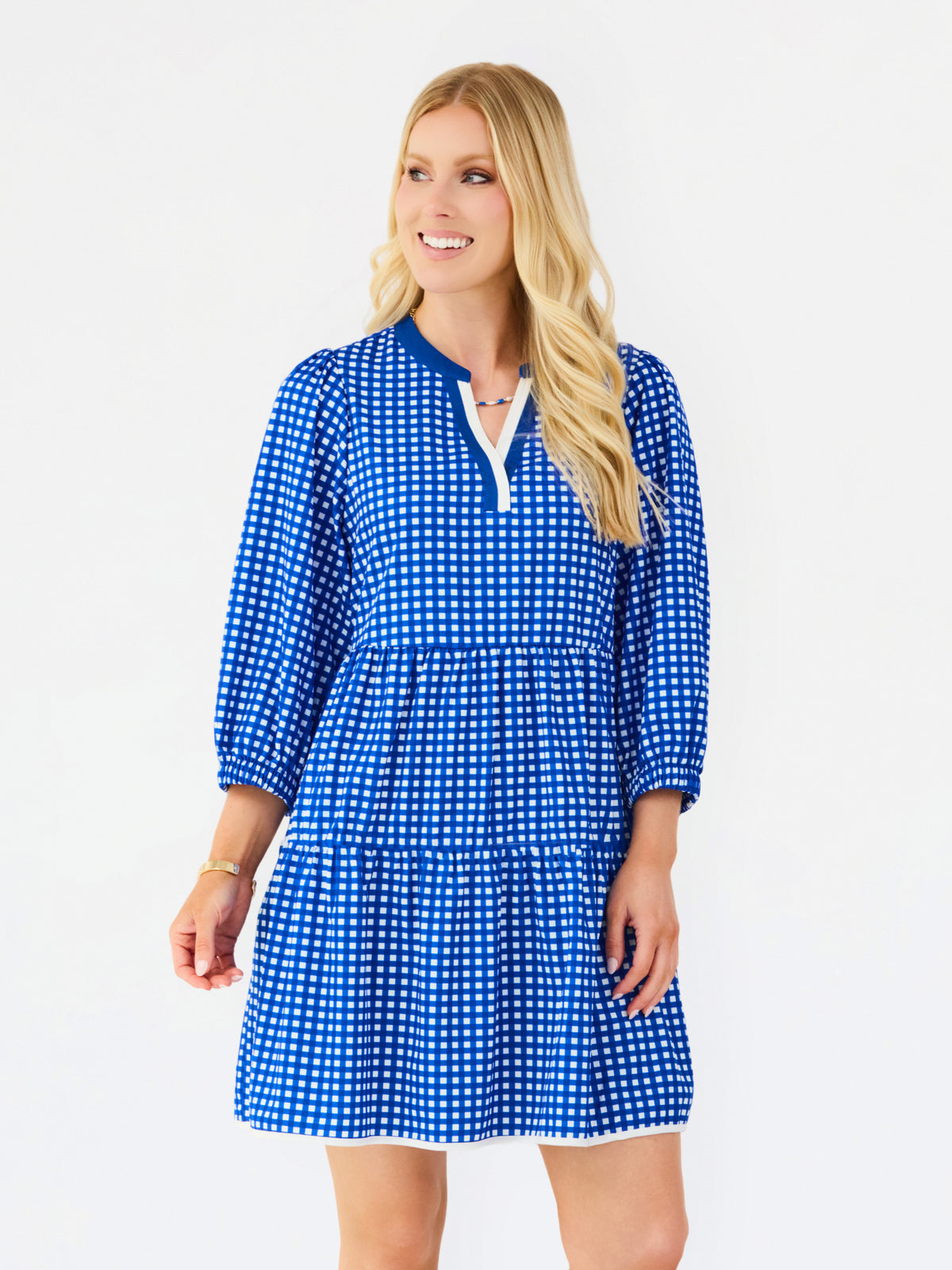 A blonde woman modeling a blue and white gingham mini dress with a split neckline and tiered skirt against a plain white studio background.