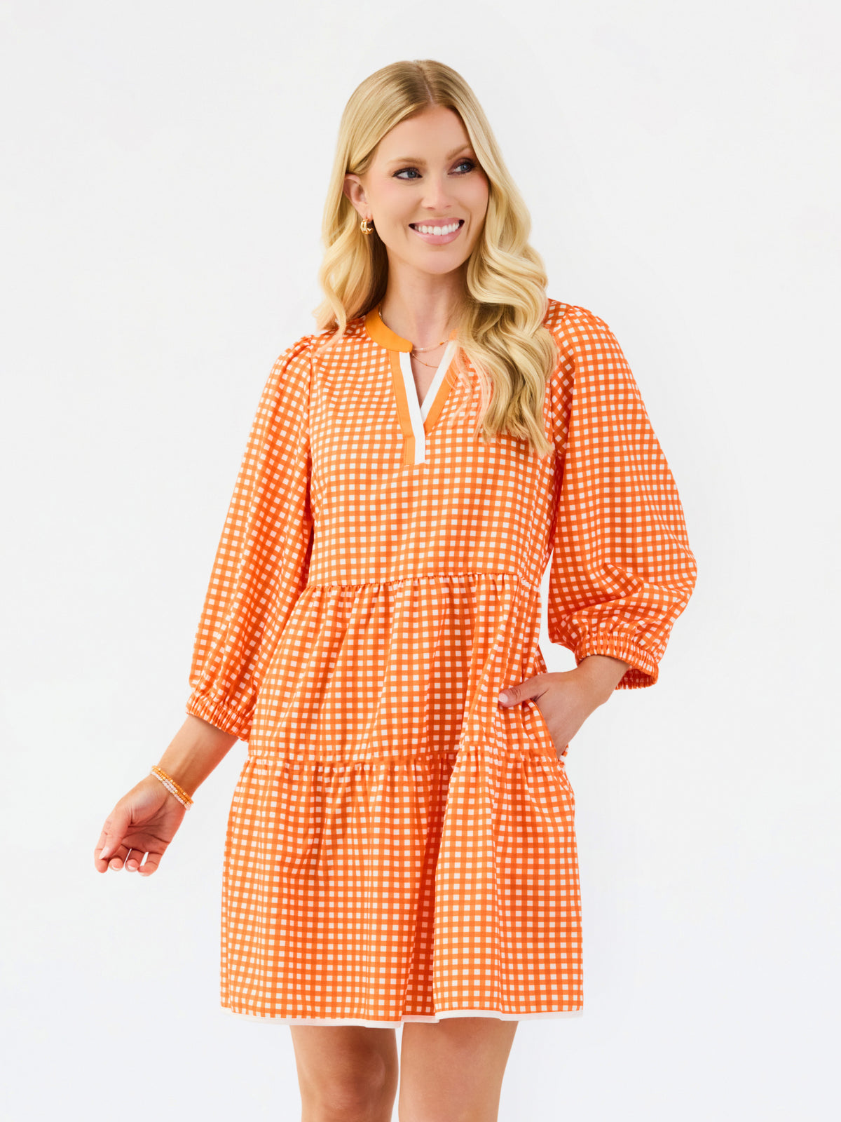 A woman modeling a tiered orange and white gingham mini dress with three-quarter sleeves and a split neckline, set against a plain white studio background.