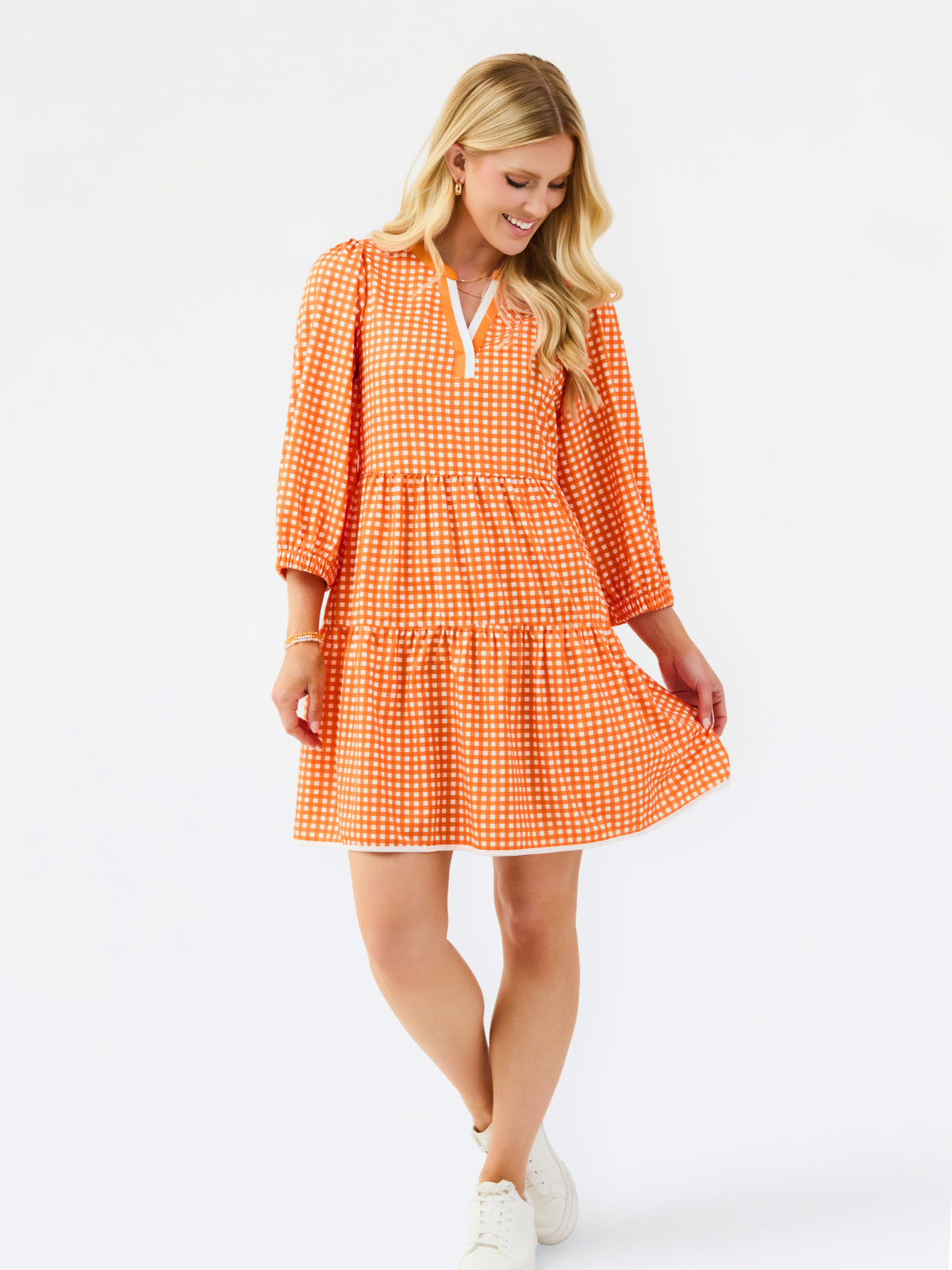 A woman modeling a tiered orange and white gingham mini dress with three-quarter sleeves and a split neckline, set against a plain white studio background.