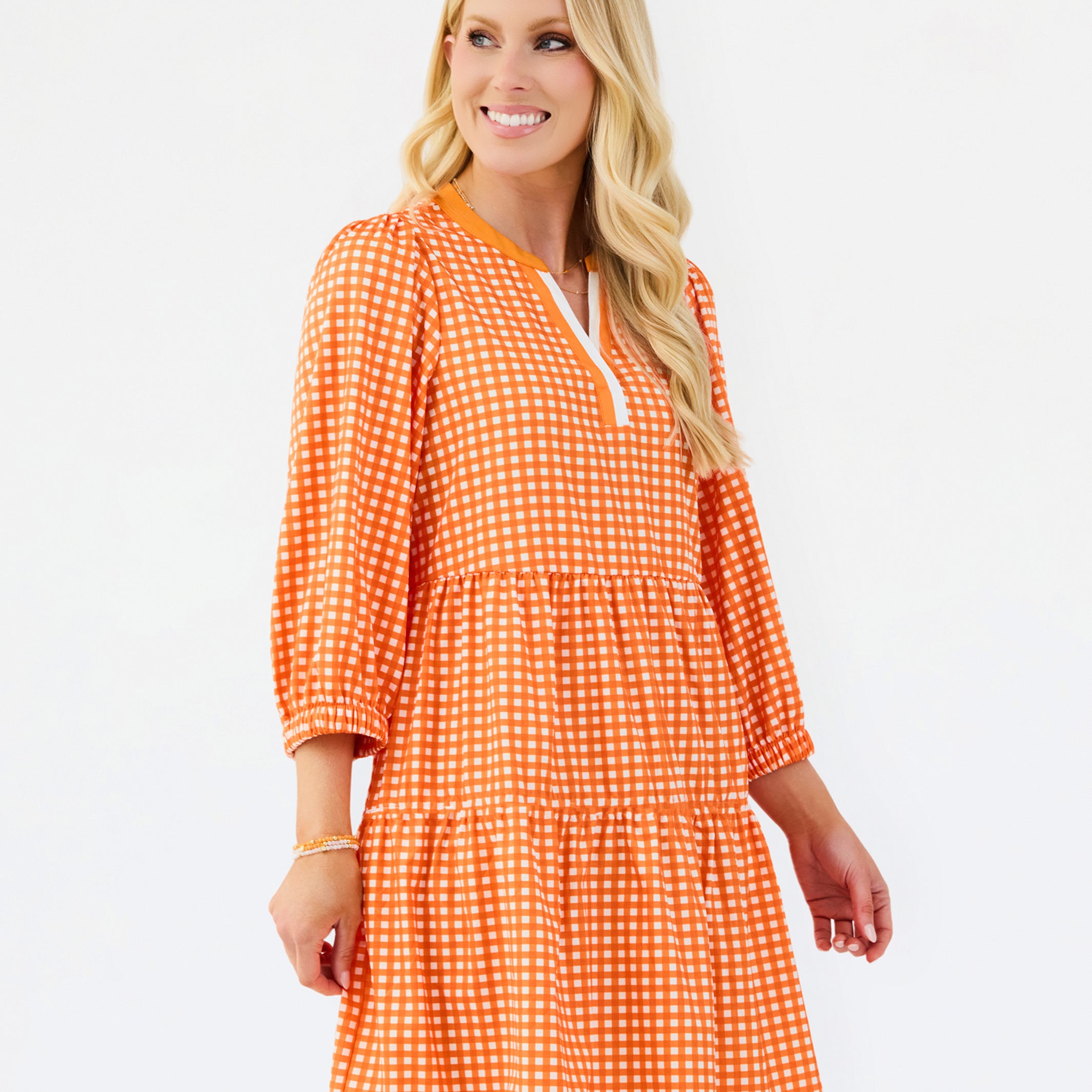 A woman modeling a tiered orange and white gingham mini dress with three-quarter sleeves and a split neckline, set against a plain white studio background.