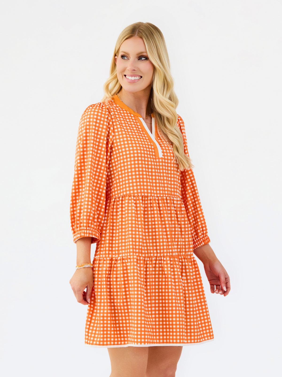 A woman modeling a tiered orange and white gingham mini dress with three-quarter sleeves and a split neckline, set against a plain white studio background.