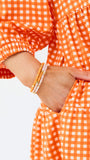 A woman modeling a tiered orange and white gingham mini dress with three-quarter sleeves and a split neckline, set against a plain white studio background.