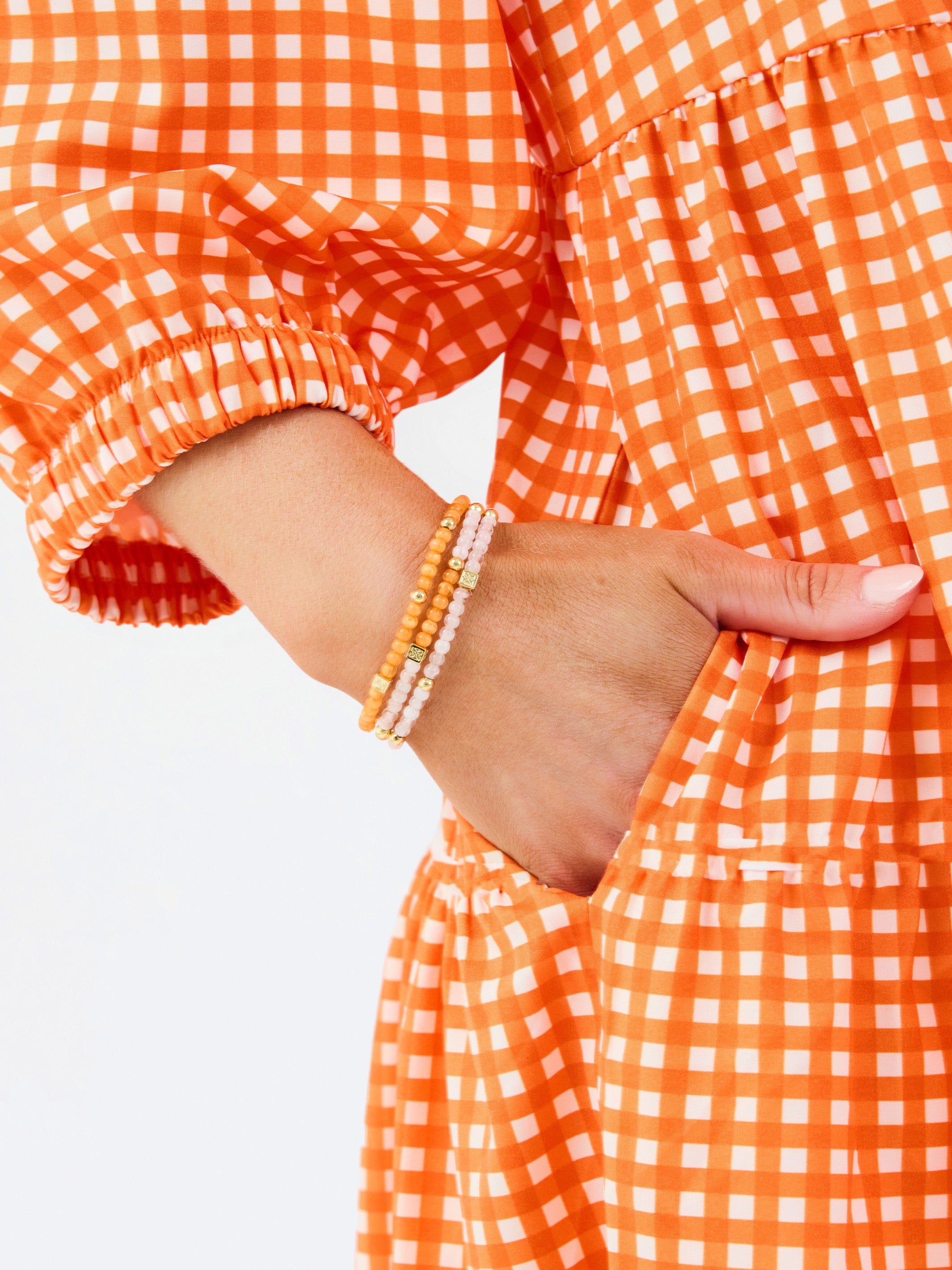 A woman modeling a tiered orange and white gingham mini dress with three-quarter sleeves and a split neckline, set against a plain white studio background.