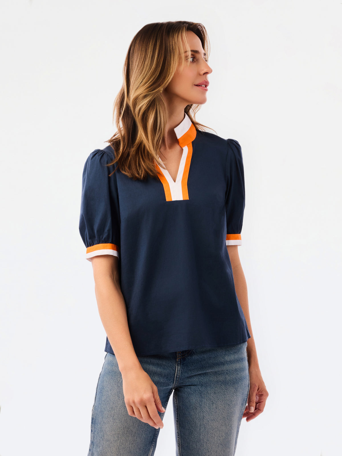 A woman modeling a navy blue short-sleeve blouse with orange and white trim on the collar and cuffs, paired with blue jeans, against a plain white studio background.