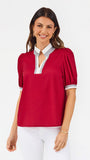 A woman modeling a short-sleeve blouse with puff sleeves, featuring a white collar and keyhole placket with red trim, paired with white pants against a light gray studio background.