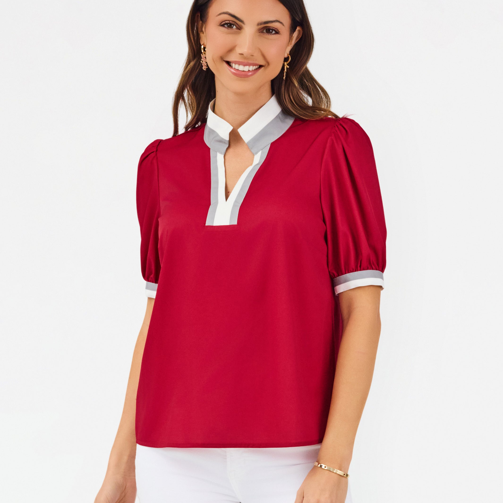 A woman modeling a short-sleeve blouse with puff sleeves, featuring a white collar and keyhole placket with red trim, paired with white pants against a light gray studio background.