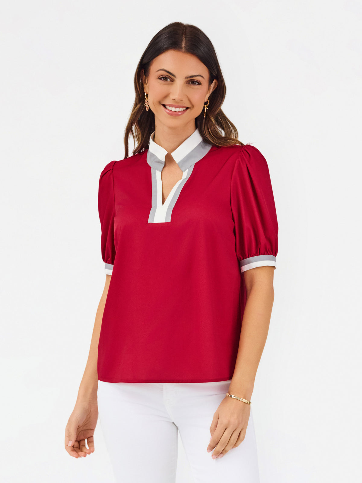 A woman modeling a short-sleeve blouse with puff sleeves, featuring a white collar and keyhole placket with red trim, paired with white pants against a light gray studio background.
