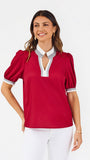 A woman modeling a short-sleeve blouse with puff sleeves, featuring a white collar and keyhole placket with red trim, paired with white pants against a light gray studio background.