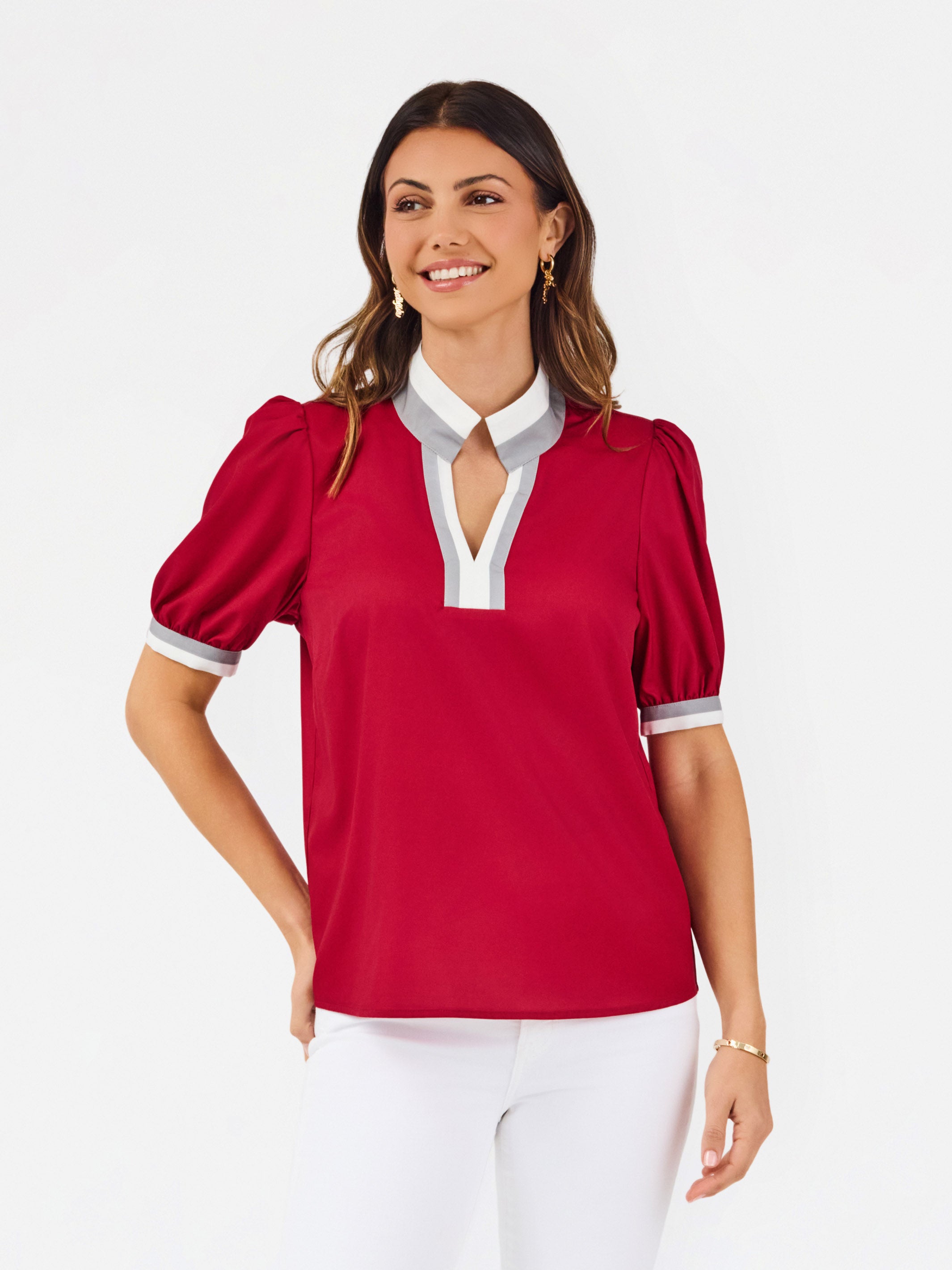 A woman modeling a short-sleeve blouse with puff sleeves, featuring a white collar and keyhole placket with red trim, paired with white pants against a light gray studio background.