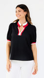 A woman modeling a black short-sleeve polo-style top with a white and red V-neck collar and matching red and white sleeve trim, paired with white pants against a light gray studio background.