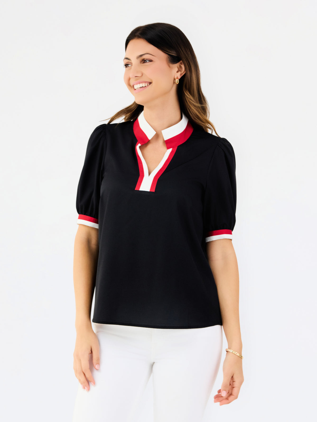 A woman modeling a black short-sleeve polo-style top with a white and red V-neck collar and matching red and white sleeve trim, paired with white pants against a light gray studio background.