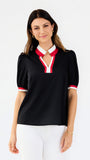 A woman modeling a black short-sleeve polo-style top with a white and red V-neck collar and matching red and white sleeve trim, paired with white pants against a light gray studio background.