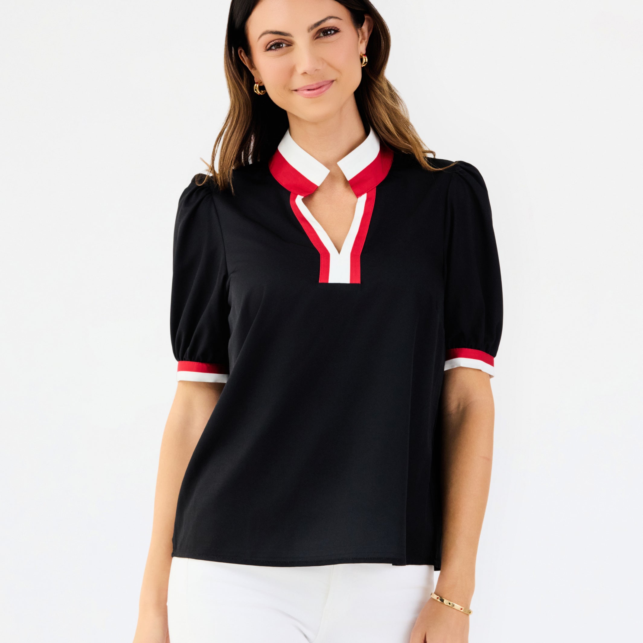 A woman modeling a black short-sleeve polo-style top with a white and red V-neck collar and matching red and white sleeve trim, paired with white pants against a light gray studio background.