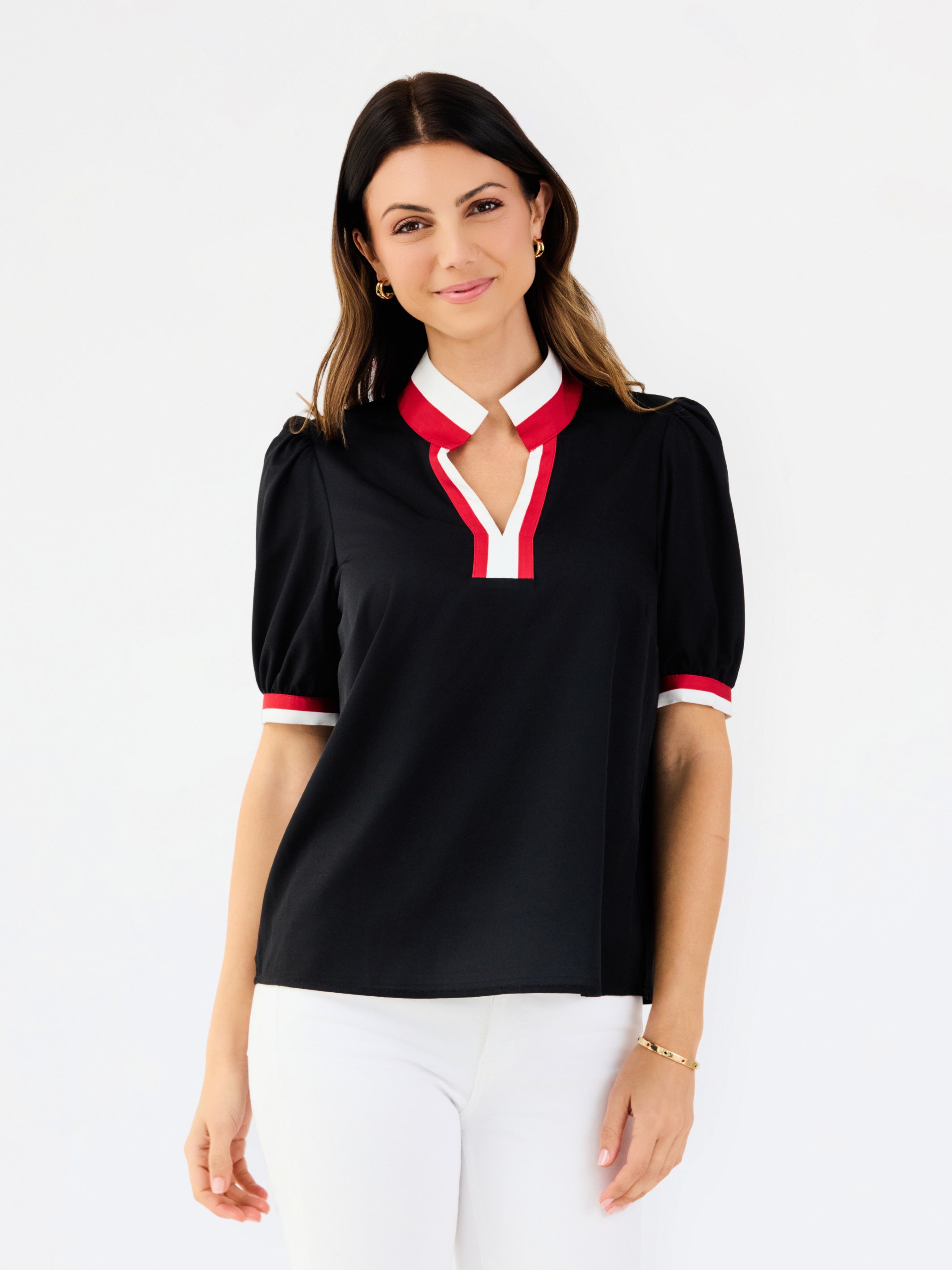 A woman modeling a black short-sleeve polo-style top with a white and red V-neck collar and matching red and white sleeve trim, paired with white pants against a light gray studio background.