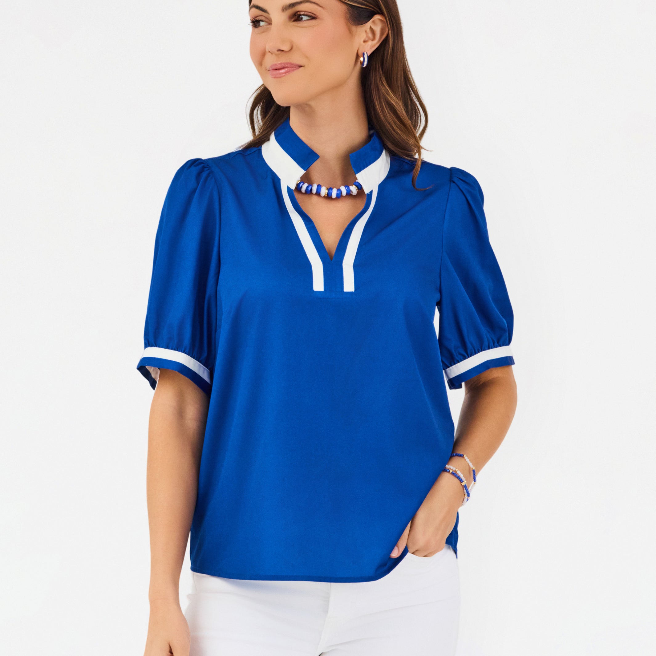 A woman modeling a royal blue short-sleeve blouse with white trim on the collar and cuffs, paired with white pants, against a plain white studio background.