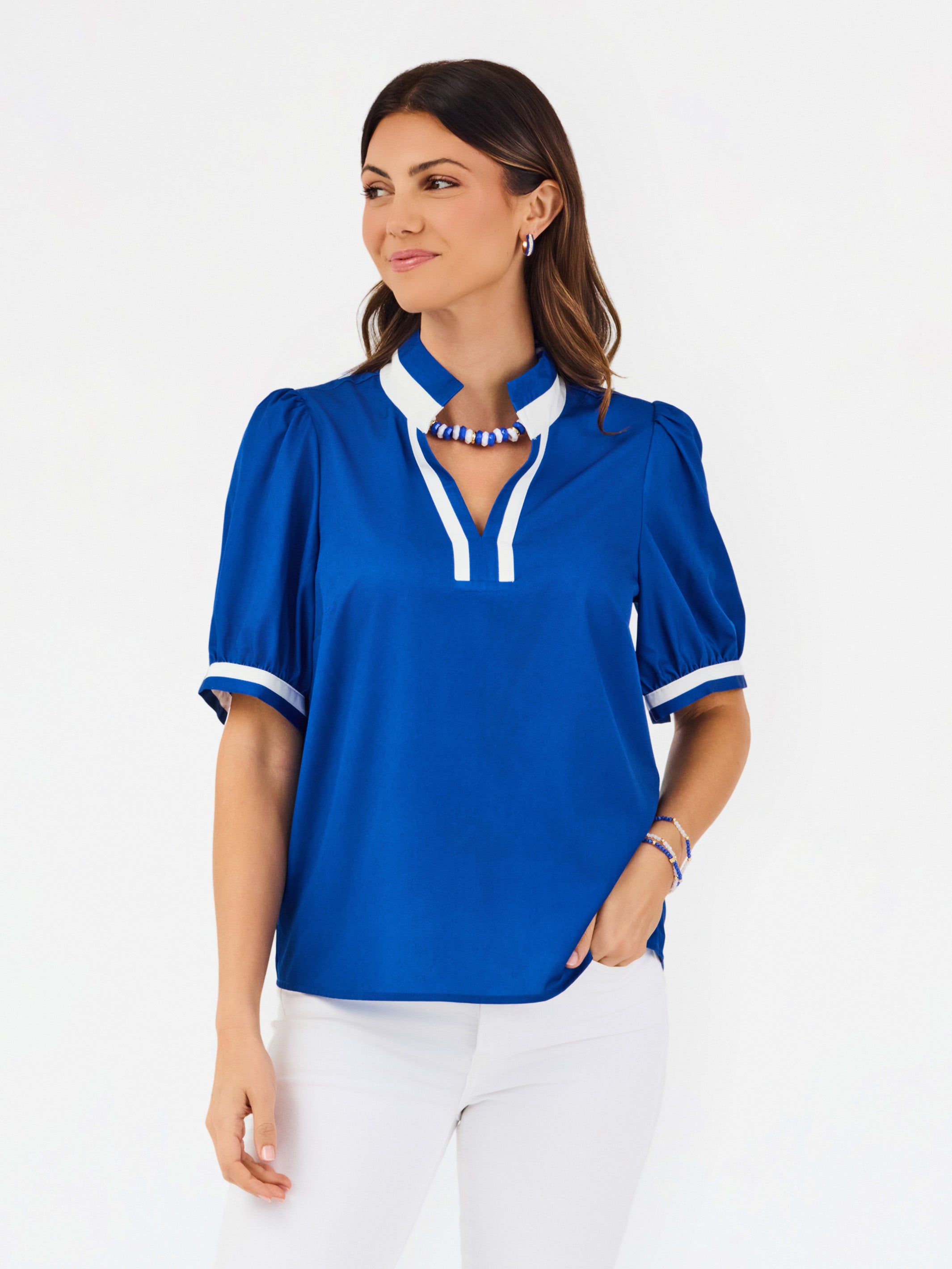 A woman modeling a royal blue short-sleeve blouse with white trim on the collar and cuffs, paired with white pants, against a plain white studio background.